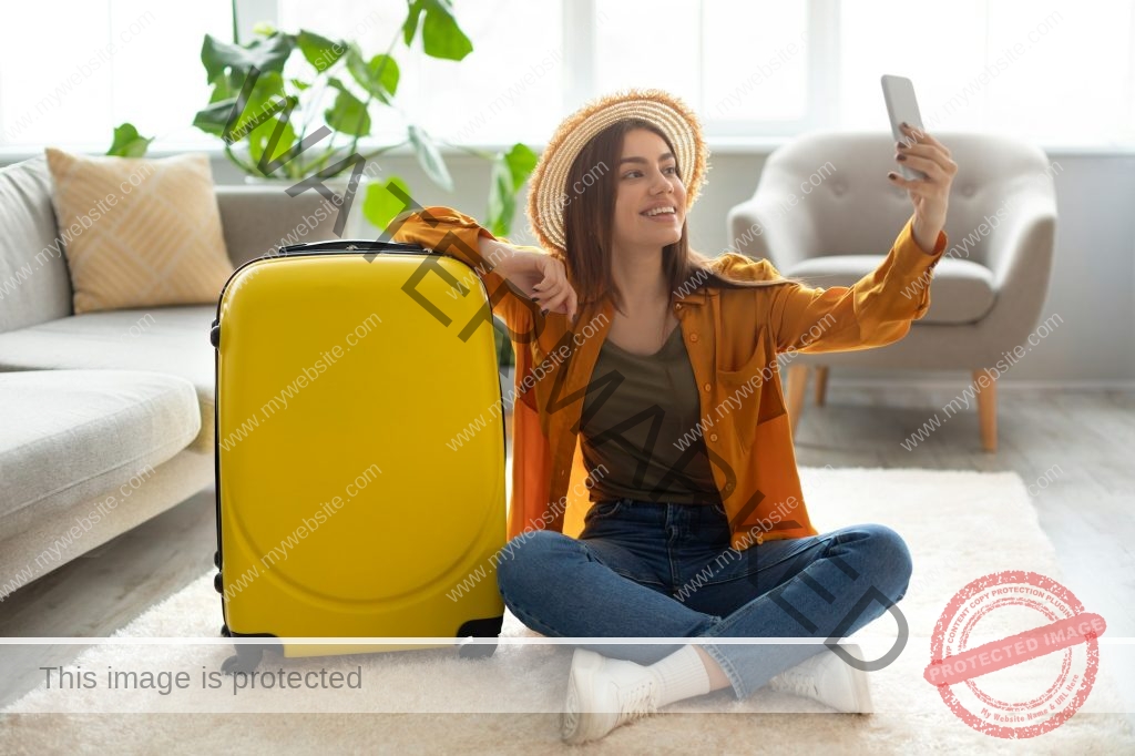 Cheerful young woman with bright suitcase taking selfie, getting ready for vacation at home