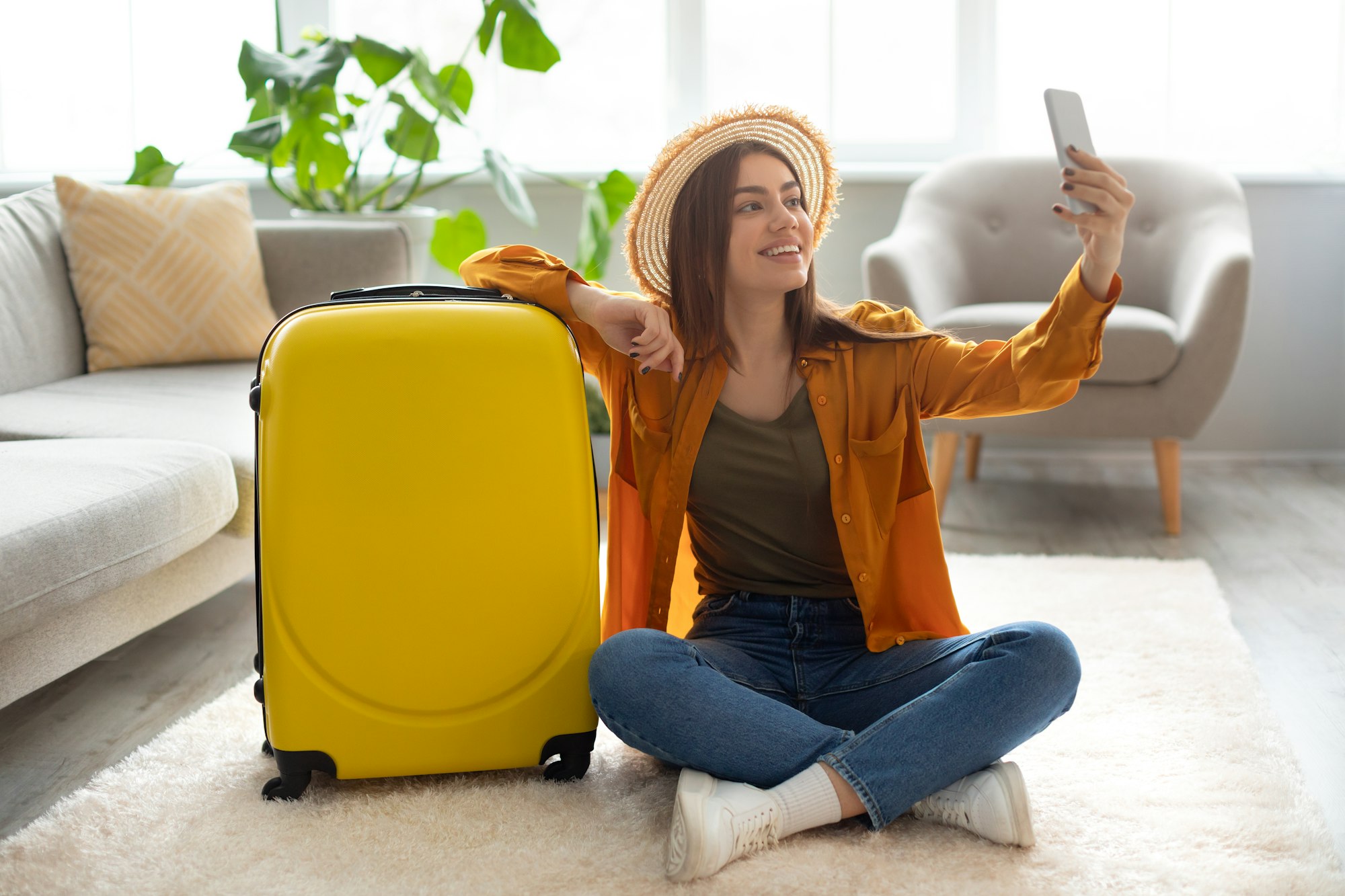 Cheerful young woman with bright suitcase taking selfie, getting ready for vacation at home